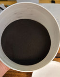 A white plastic jar filled with dark brown powder, held by a hand over a wooden desk with a white lid placed beside it.