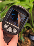 A handheld black infrared thermometer with a digital display held by a hand, showing temperature readings, with a blurred background of green plants. Caleigh Cassidy Lanthier Photography 2026